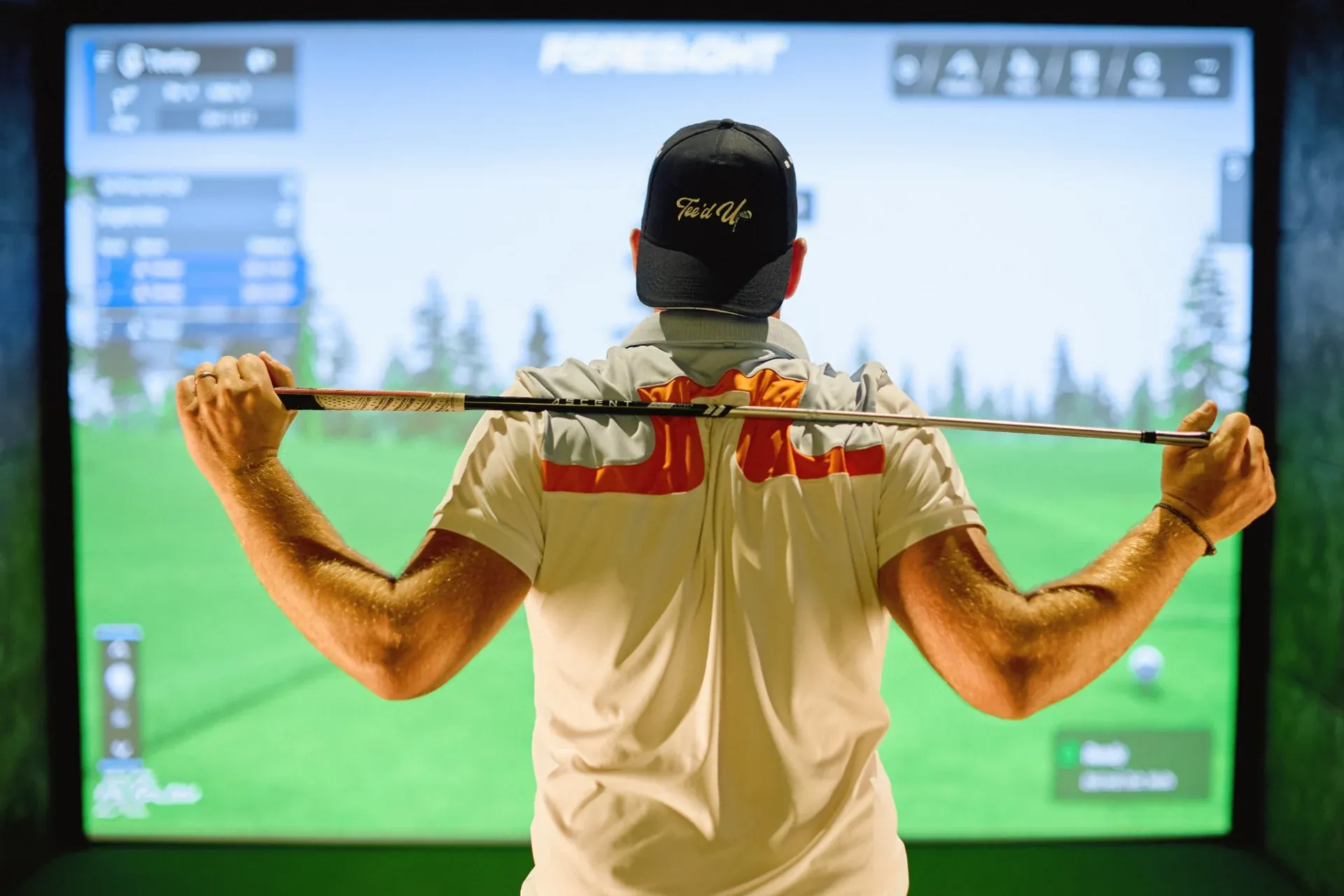 A golfer wearing a Tee’d Up cap in front of an indoor golf simulator